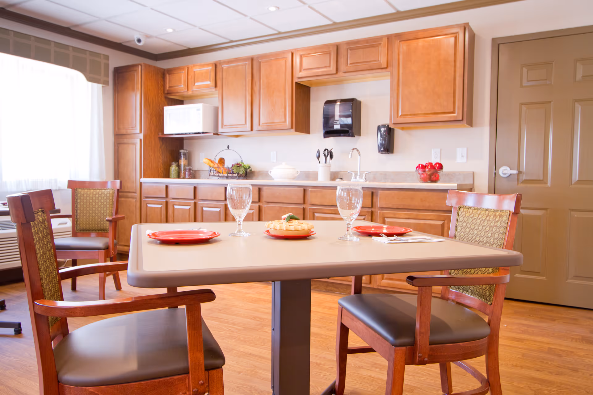 Dining area with a table and chairs set with plates and glasses in front of a kitchenette with wooden cabinets.