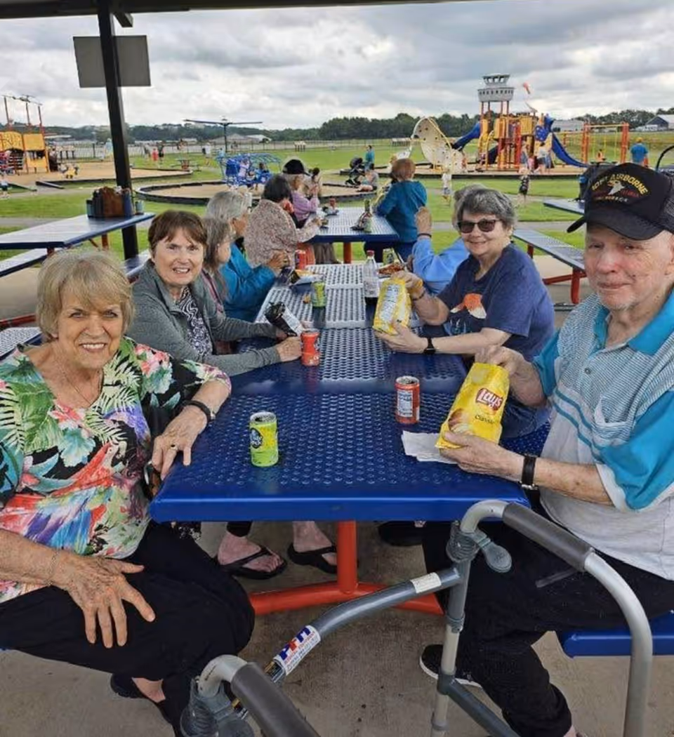 A group of elderly people sitting at blue picnic tables under a covered pavilion, enjoying snacks and drinks. In the background, there is a playground with slides and climbing structures, and an open grassy area under a cloudy sky.