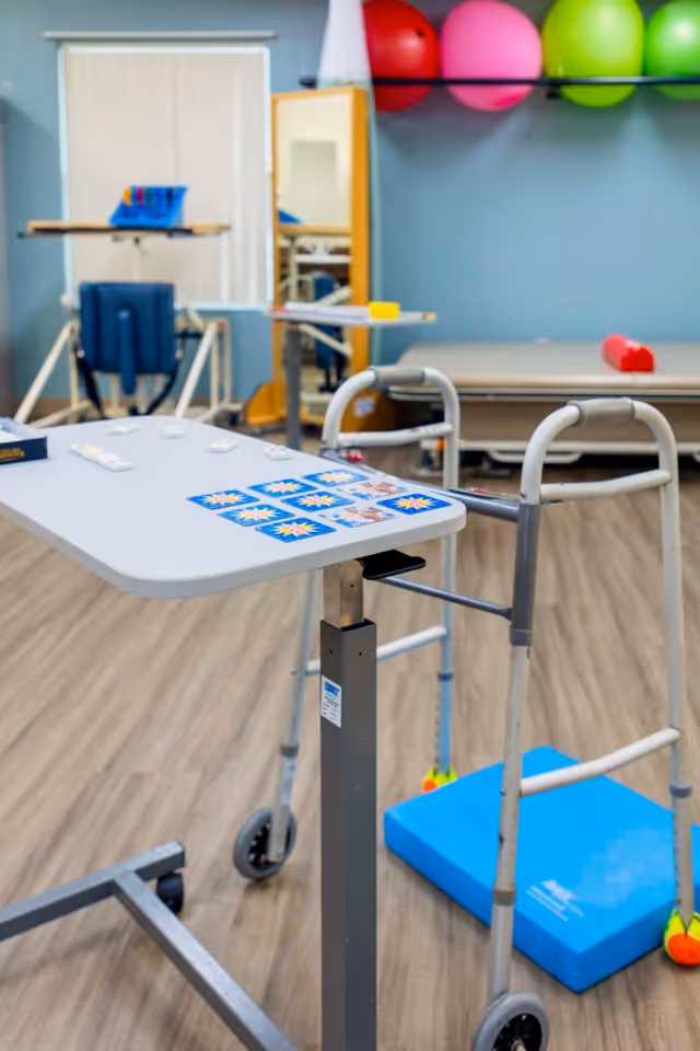 Therapy room with a walker, adjustable over-table, exercise equipment and colorful therapy balls on a shelf.