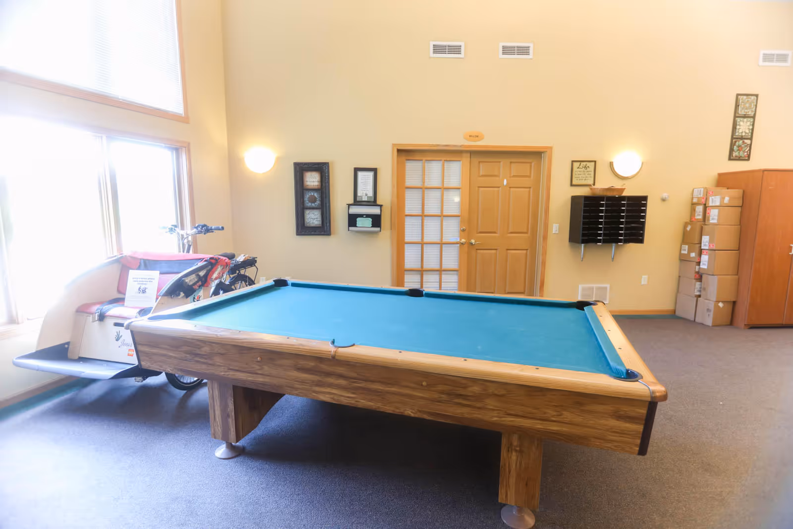 A bright room with a pool table in the center, a large window on the left letting in natural light, a mobility scooter parked near the window, wall-mounted mailboxes, framed pictures, and a wooden door with a glass panel.