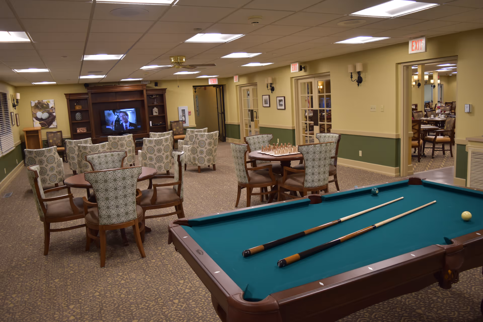 A recreational room in Sedgwick Plaza featuring a pool table with two cues and balls, several round tables with patterned chairs, a chess set on one table, and a seating area with armchairs facing a television mounted on a wooden entertainment center. The room has beige walls with green wainscoting, carpeted floor, ceiling lights, and multiple exit signs. A dining area is visible through an open doorway.