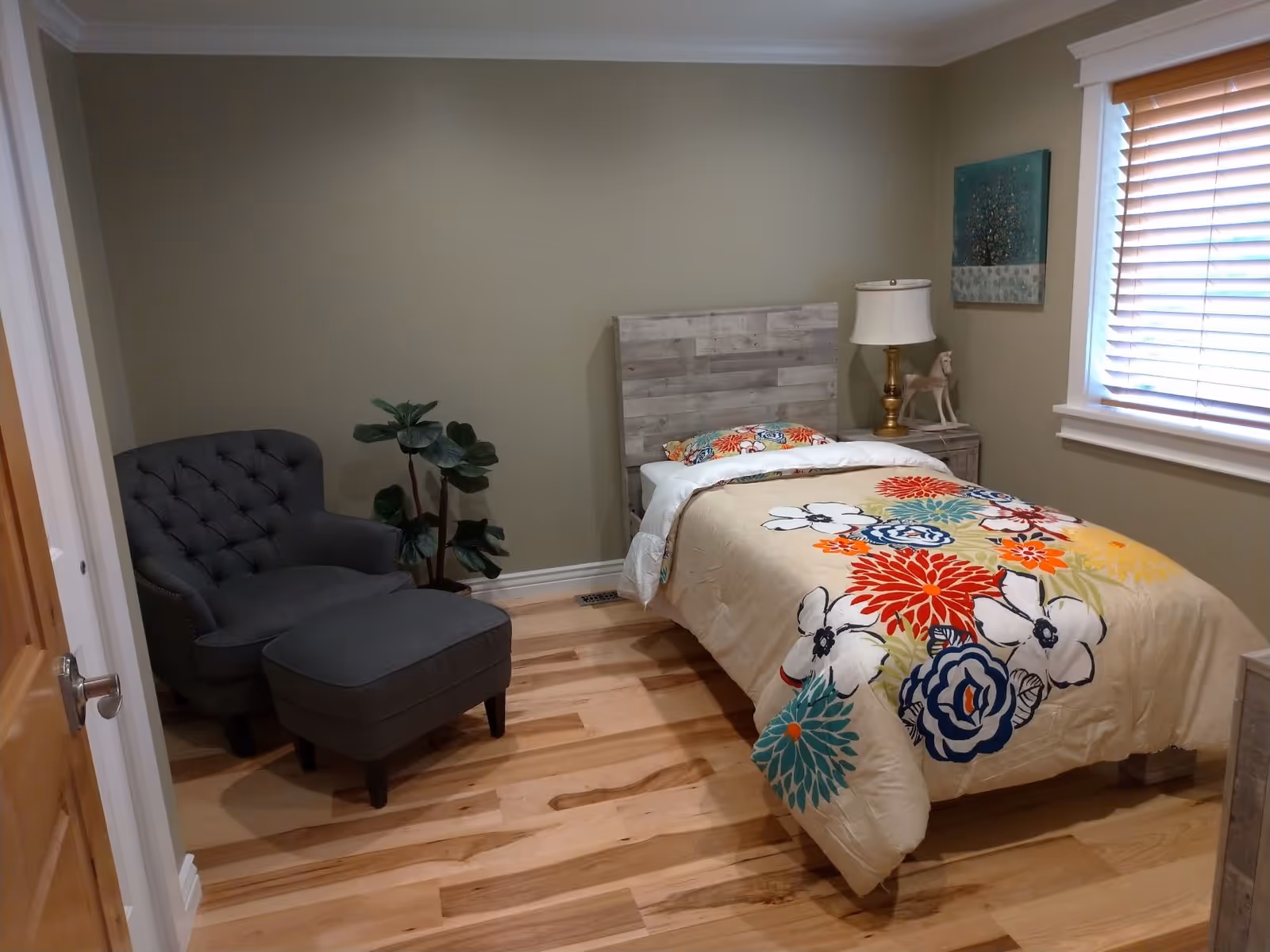 Small bedroom with a single bed featuring a colorful floral comforter, a gray tufted armchair with ottoman, wooden floors and a window with blinds.