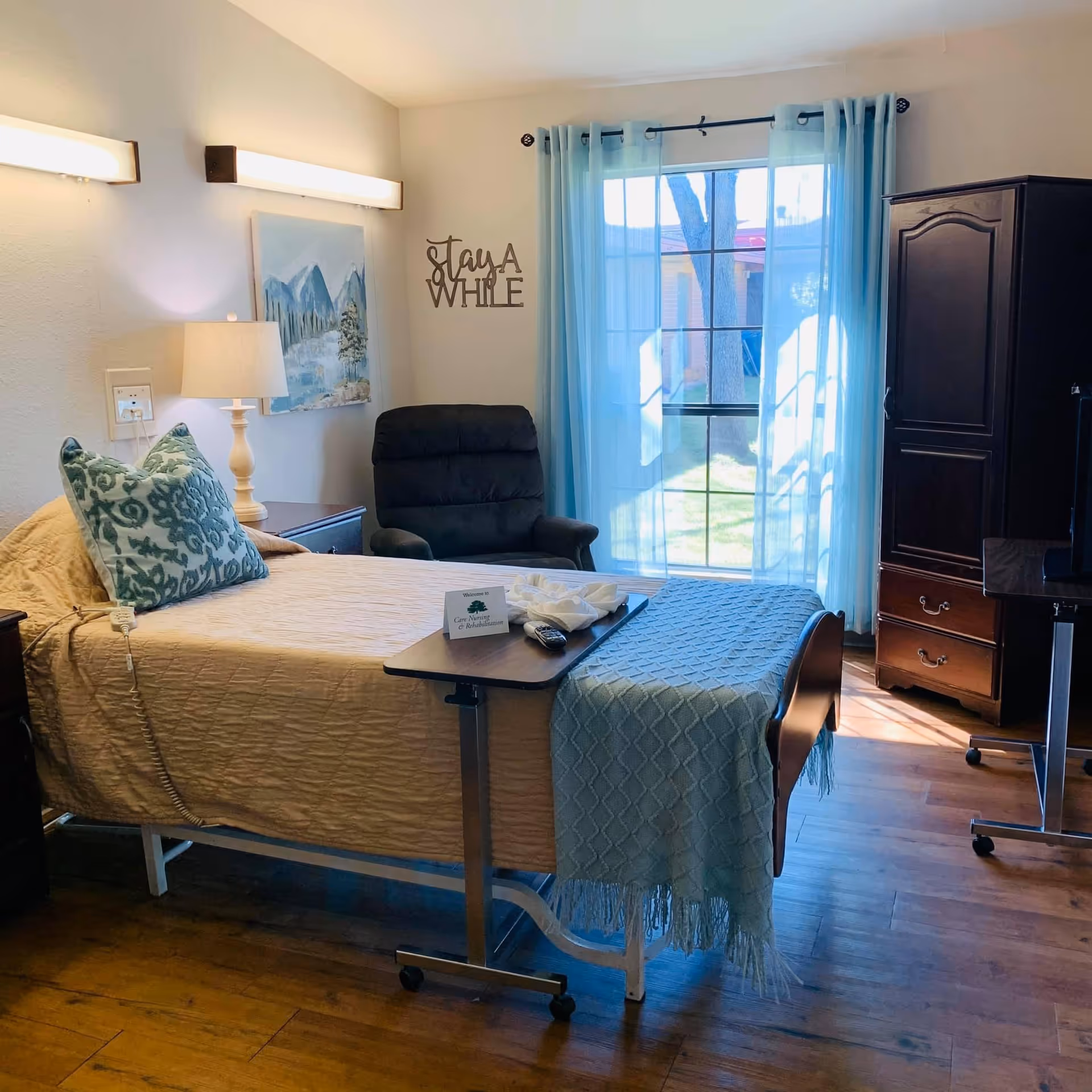 A cozy bedroom in a nursing and rehabilitation facility featuring a single bed with beige bedding and a blue patterned pillow. A small overbed table holds a remote control and a welcome card. There is a black recliner chair next to the bed, a wooden armoire, and a TV on a stand. Light blue curtains cover a large window letting in natural light. Wall decor includes a painting of mountains and a sign that says 'Stay A While'.