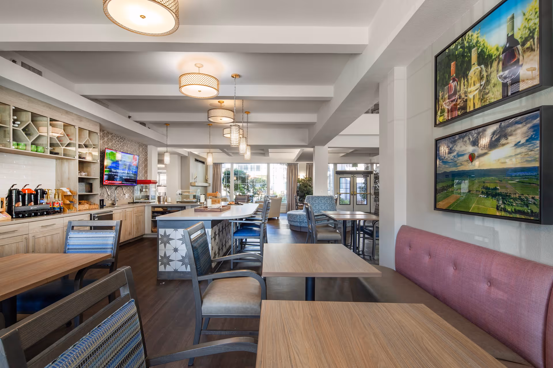 A bright and modern dining area in Oakmont Gardens featuring wooden tables and chairs, a long counter with coffee and snack stations, pendant lighting, and large windows letting in natural light. The walls are decorated with framed landscape and wine-themed pictures.