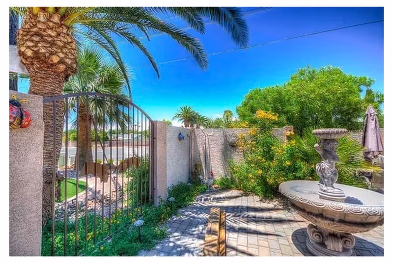 Outdoor patio area with a stone birdbath fountain, surrounded by green plants and trees including palm trees. There is a metal gate on the left side and a clear blue sky above.
