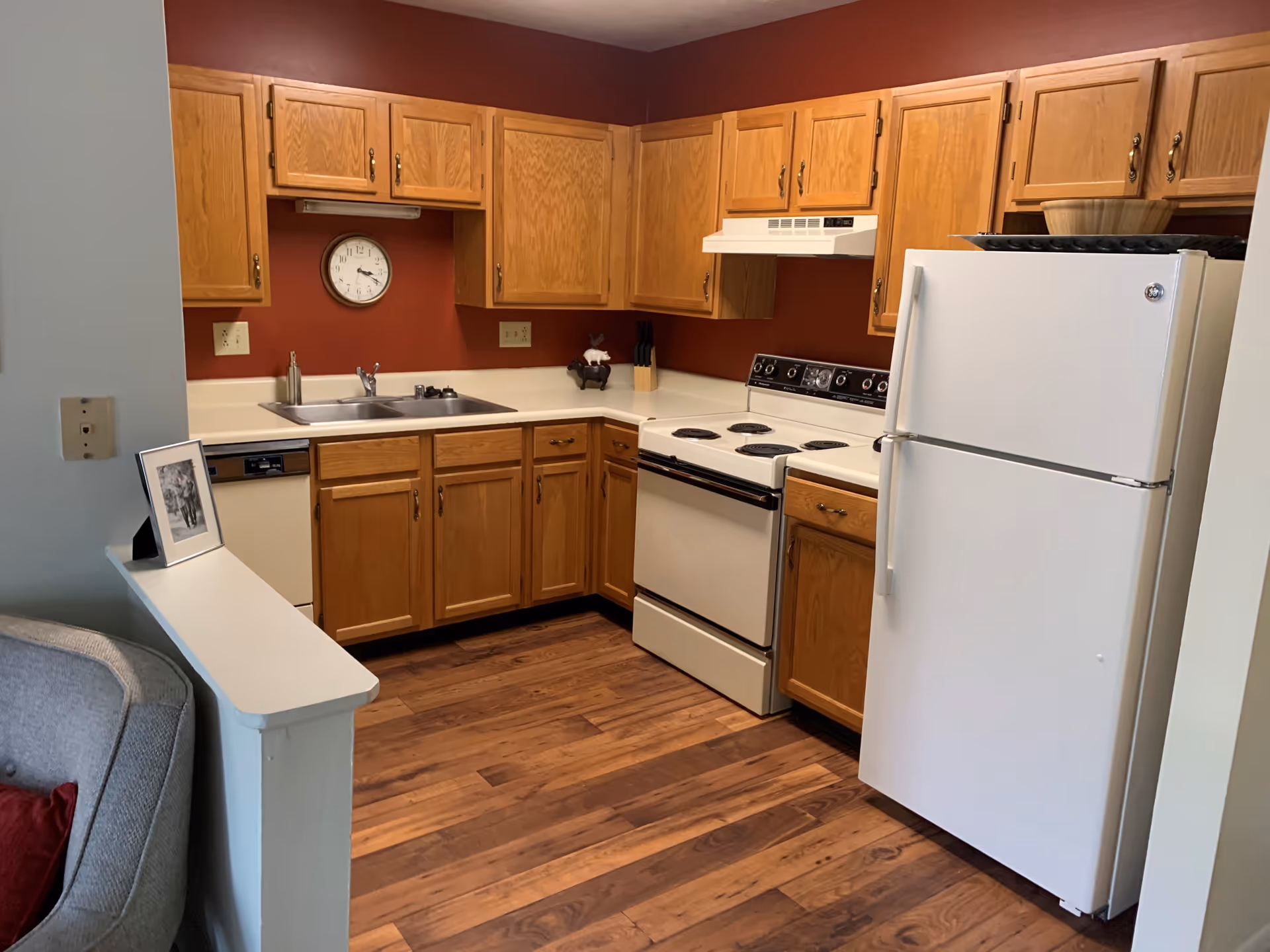A kitchen with wooden cabinets, white countertops, a white refrigerator, an electric stove with four burners, a double sink, and a dishwasher. The walls are painted a deep red color, and the floor has a wood-like finish. A clock is mounted on the wall above the sink, and a small framed photo is placed on a half wall next to a gray upholstered chair.