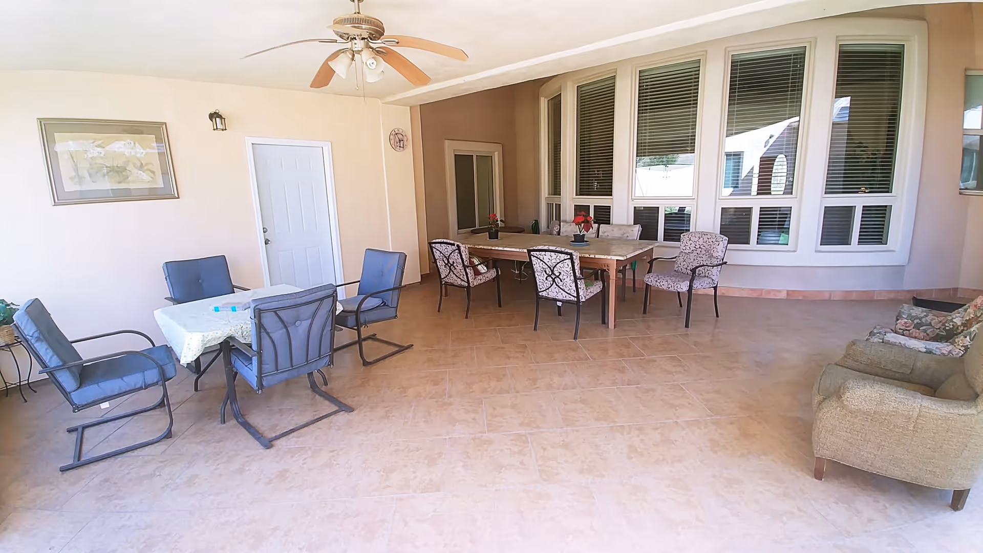 Covered outdoor patio area with tiled floor, featuring two seating areas. One area has a small table with a white tablecloth and four blue cushioned chairs. The other area has a larger wooden table with six patterned cushioned chairs. There is a ceiling fan above and a framed picture on the wall near a white door. Large windows with blinds are visible on the right side.