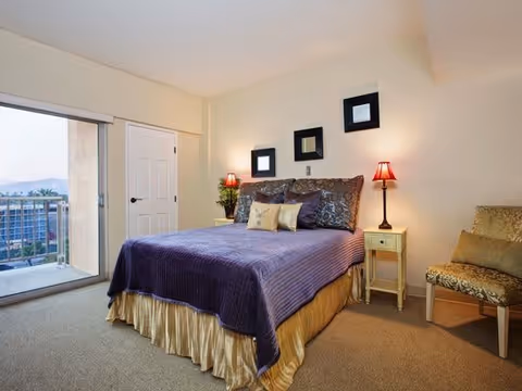 A bedroom with a large bed covered in a purple blanket and multiple pillows. There are two bedside tables with red lamps on each side of the bed. Three small square mirrors are hung on the wall above the bed. A cushioned chair is placed to the right side of the bed. A sliding glass door on the left side opens to a balcony with a view of buildings and mountains in the distance.