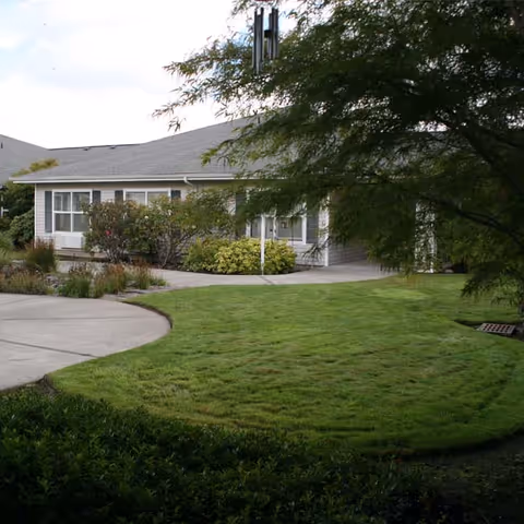 Single-story memory care building front with a manicured lawn, curved concrete walkway, shrubs, and a tree partially obscuring the view.