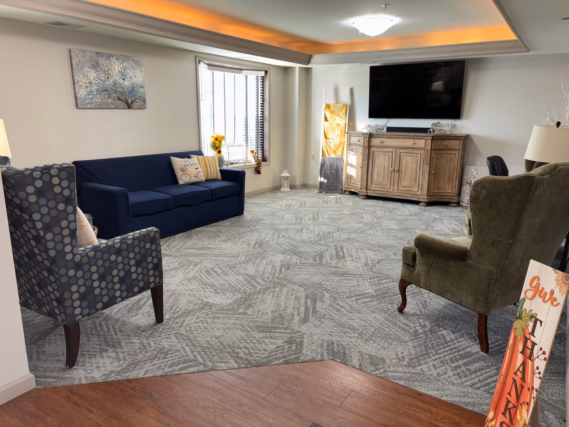 A cozy living room featuring a blue sofa, patterned armchair, and a wooden cabinet with a TV. Sunlight streams through a window adorned with yellow flowers, creating a warm atmosphere.