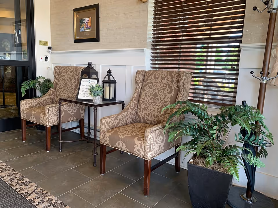 A waiting area with two patterned armchairs separated by a small table holding two decorative lanterns and a small potted plant. There is a large potted fern plant on the floor next to the right chair. Behind the chairs is a window with wooden blinds, and to the left is a glass door with a keypad entry system. A coat rack with umbrellas is visible on the right side.
