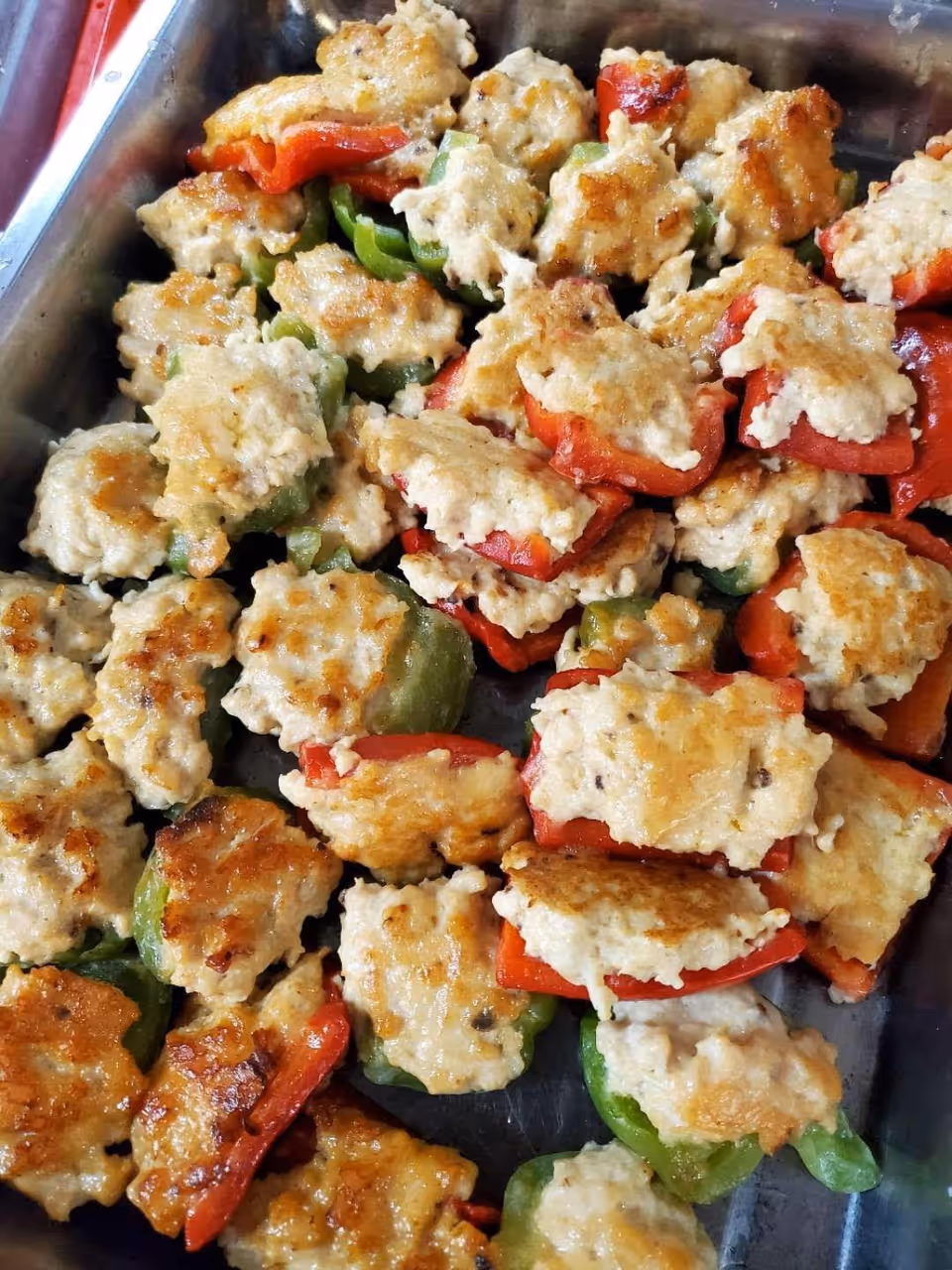 A tray filled with cooked stuffed bell pepper halves, with a browned topping that appears to be a cheese or meat mixture, featuring both red and green bell peppers.