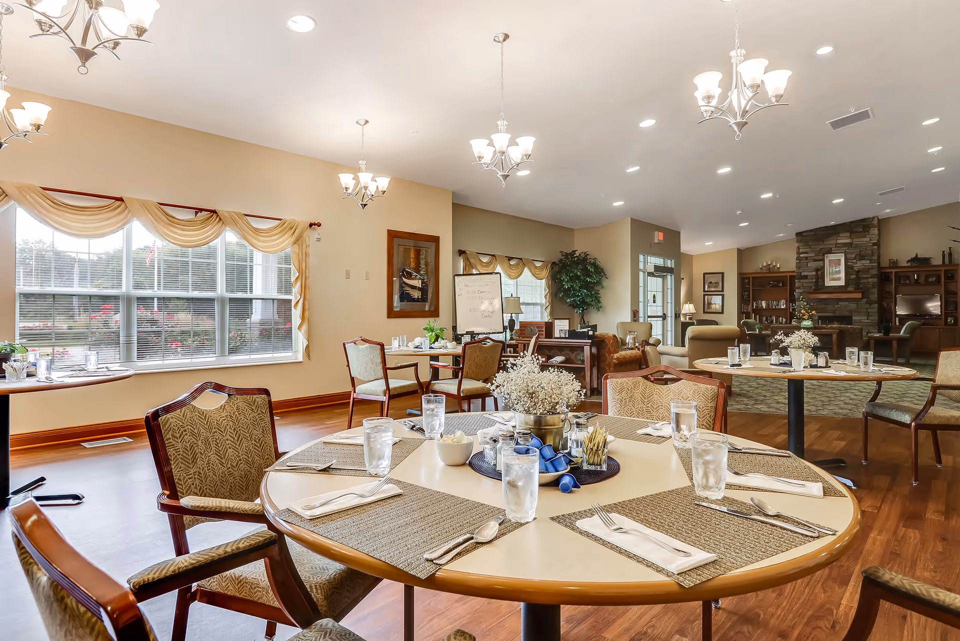 A bright and spacious dining room in an assisted living facility with round tables set with placemats, utensils, glasses of water, and a centerpiece of white flowers. The room features large windows with beige curtains, wooden flooring, multiple chandeliers, and a cozy seating area with armchairs near a stone fireplace in the background.