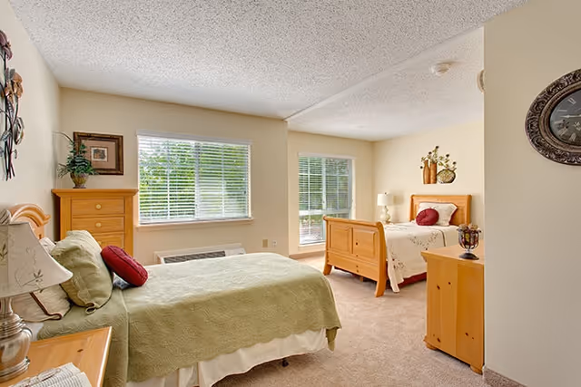 Bright shared bedroom with two wooden twin beds, matching dressers, and large windows with blinds.