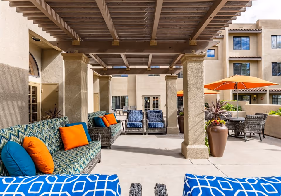 Outdoor patio area at Fairwinds - Rio Rancho featuring cushioned wicker seating with colorful blue and orange pillows under a wooden pergola, large potted plants, and tables with orange umbrellas. The building exterior is visible in the background.