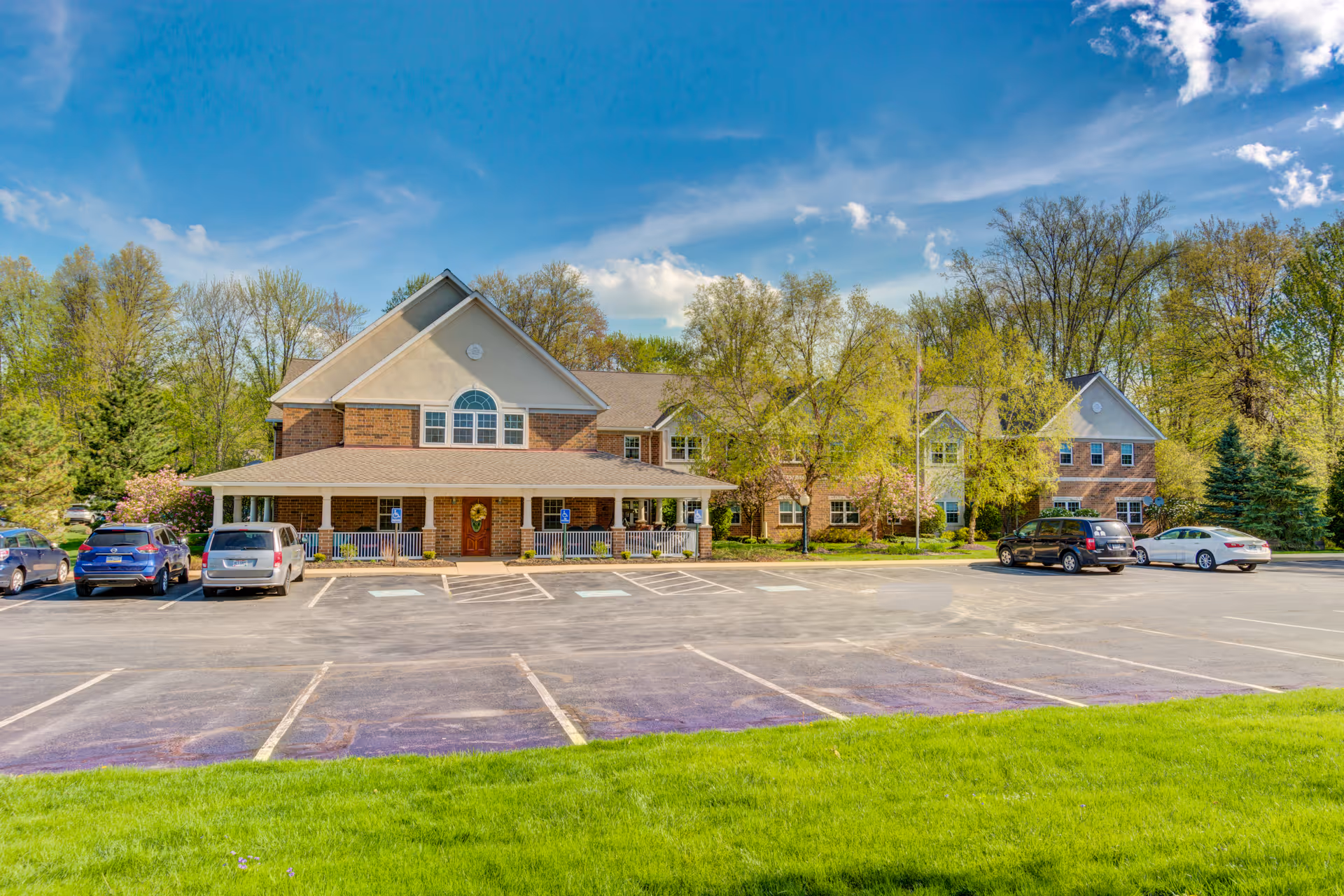 Exterior view of Symphony at Olmsted Falls facility showing a large brick building with a covered porch, surrounded by trees and a parking lot with several parked cars under a blue sky with some clouds.