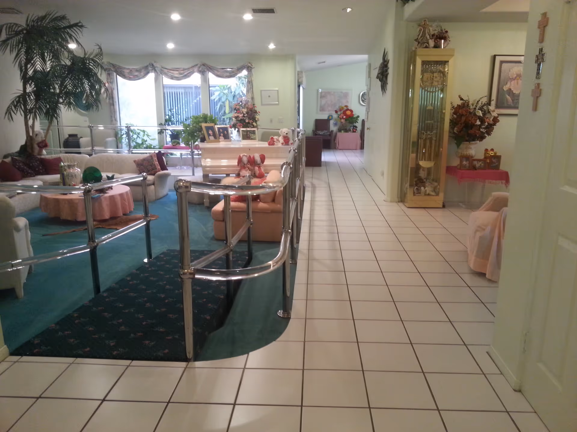 Interior view of a senior living facility showing a living room area with a white sectional sofa, a round table with a pink tablecloth, plants, framed photos on a white piano, and a hallway with tiled floor leading to other rooms. The living room area is carpeted in green and has chrome handrails around a small ramp. There are decorative items including flowers, a grandfather clock, and wall art.