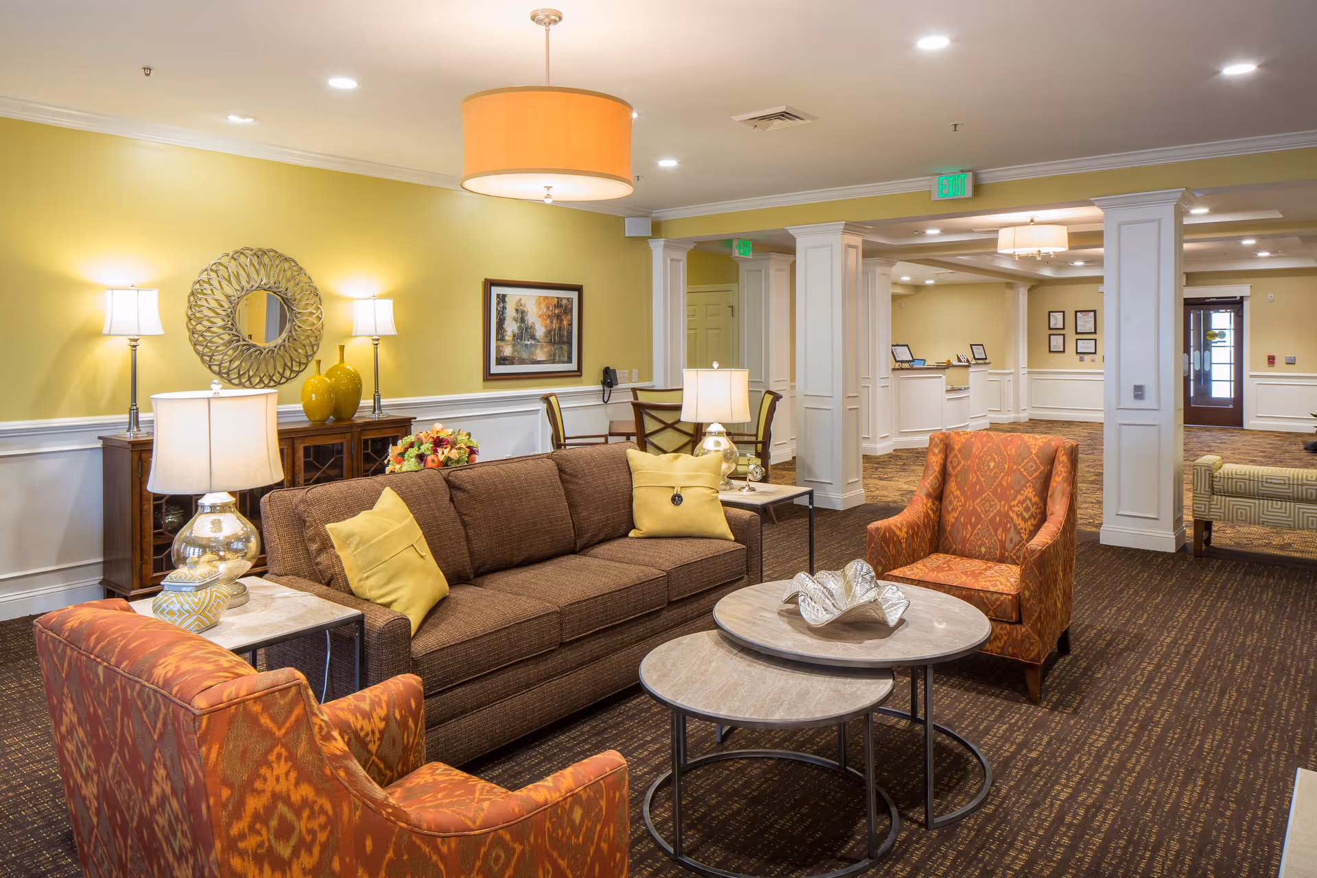 Well-lit senior living lounge with a brown sofa, two patterned armchairs, nested coffee tables and lamps in front of a yellow wall and reception area in the background.
