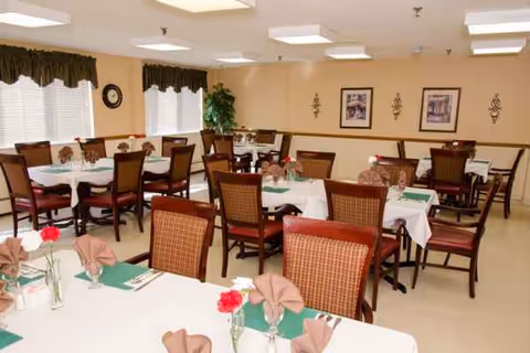 A bright dining room with round tables, brown chairs, and green napkins in a senior facility.