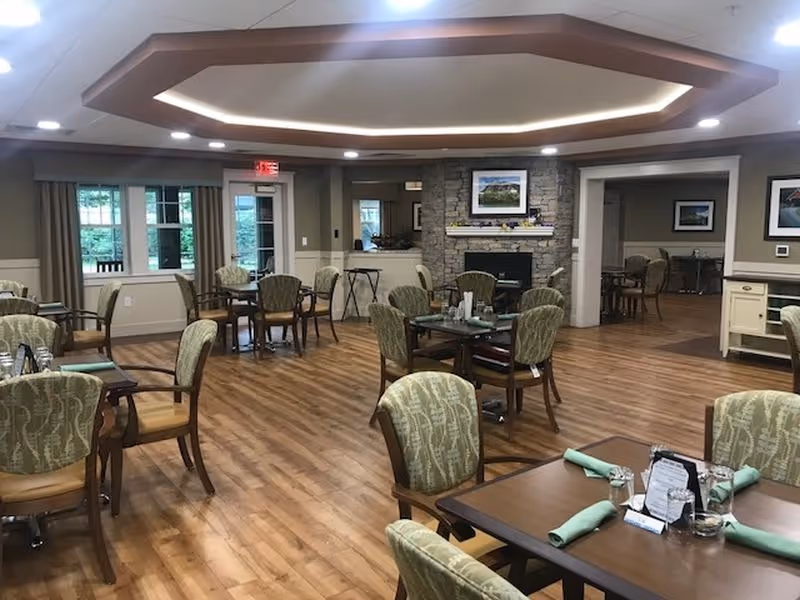 A spacious dining room with multiple wooden tables and cushioned chairs arranged neatly. The room features wood flooring, a stone fireplace with a framed picture above it, and large windows with curtains allowing natural light. The ceiling has a decorative recessed design with lighting. Tables are set with green napkins and glassware.