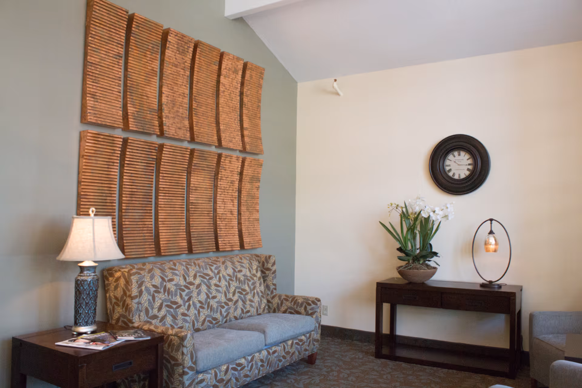 A cozy living room area with a patterned sofa, a side table with a lamp and magazines, a decorative wall art made of wooden panels, a console table with a potted plant and a modern lamp, and a round wall clock on a beige wall.