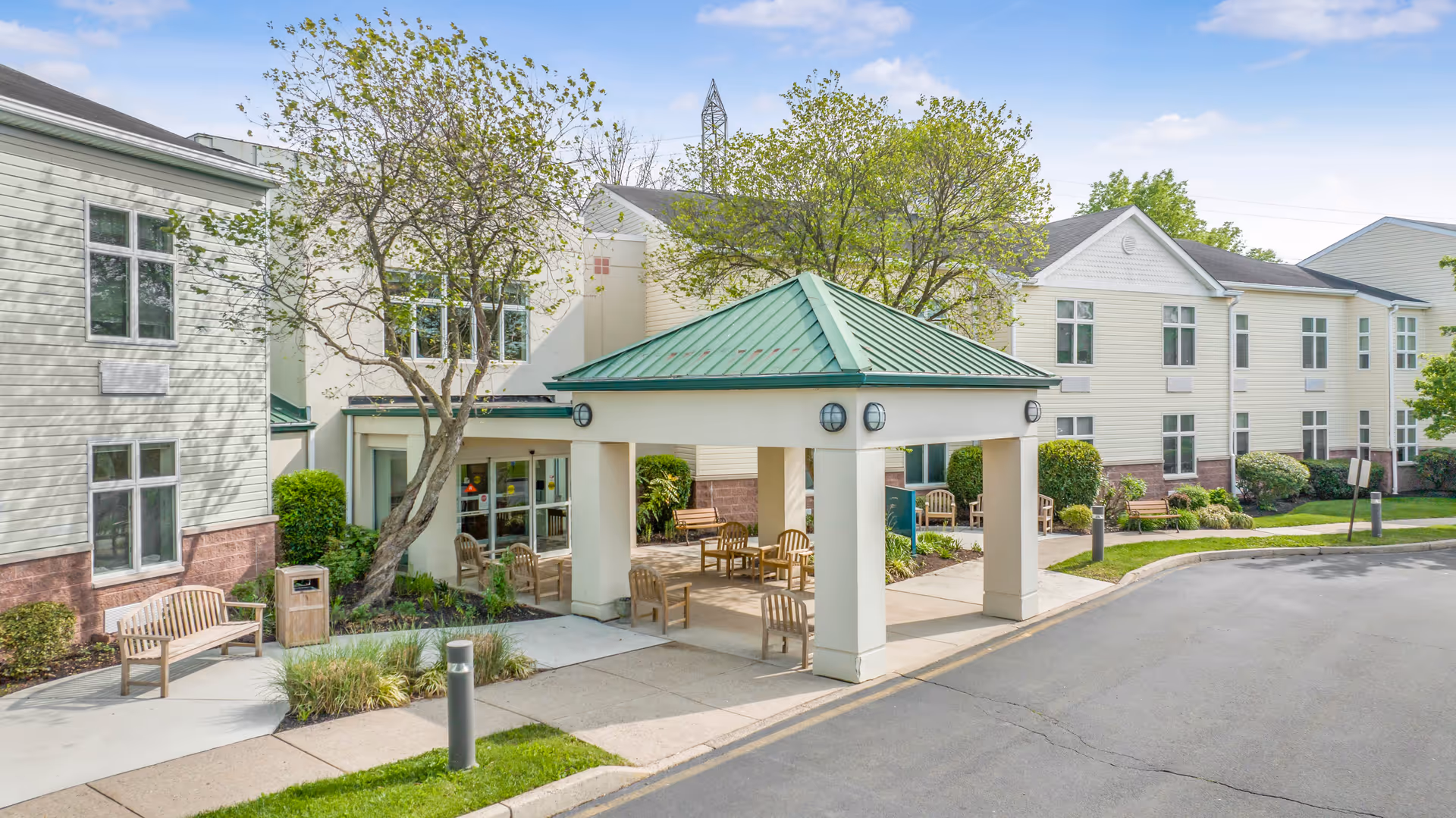 Exterior view of The Terraces at Lawrence senior living facility showing a covered entrance with a green roof, several wooden benches, trees, shrubs, and a paved driveway under a partly cloudy sky.