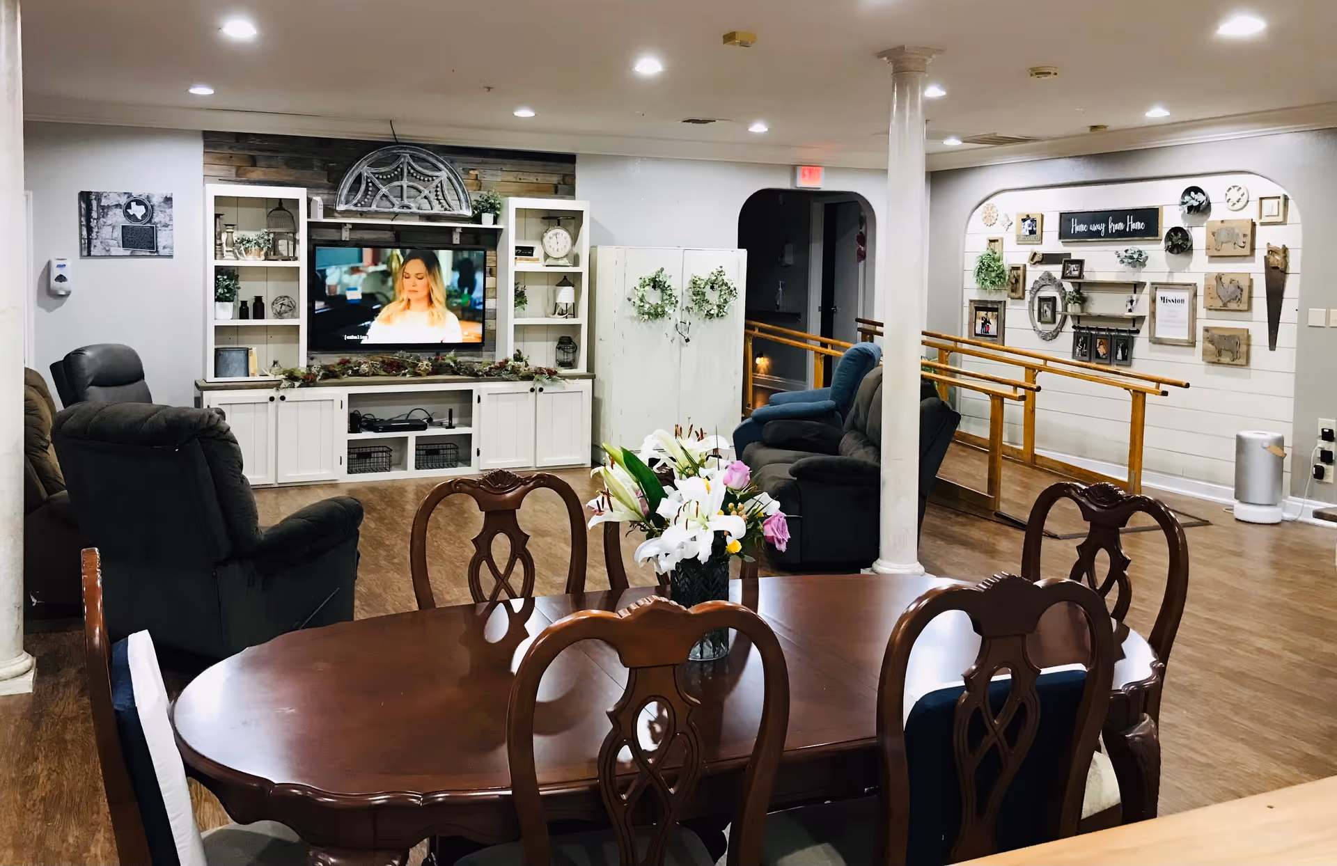 A cozy assisted living common area with a wooden dining table and chairs in the foreground, a vase with flowers on the table, several recliner chairs facing a wall-mounted TV, and decorative shelves and wall art in the background. The room has wooden flooring, white walls, and a handrail along a ramp near the back wall.