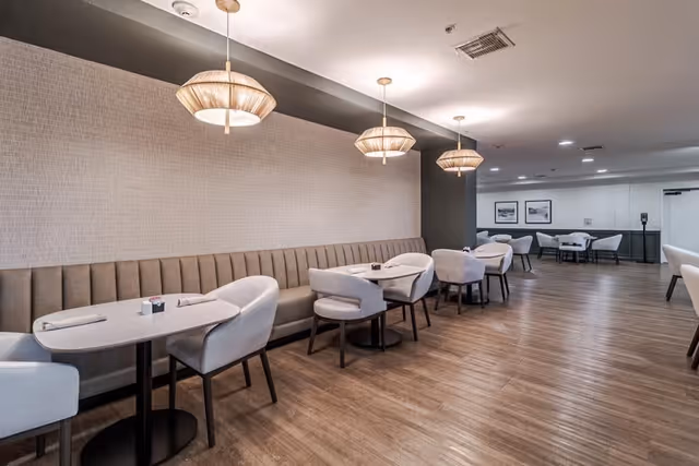 A modern dining area with multiple tables and cushioned chairs arranged along a long upholstered bench against a textured wall. Three pendant lights hang from the ceiling, illuminating the space with wooden flooring and framed pictures on the far wall.