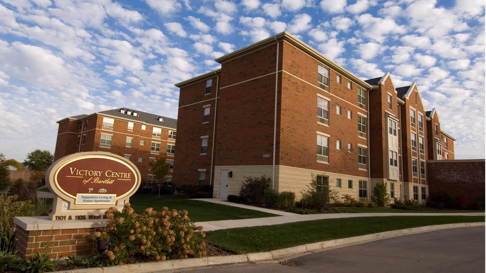 Exterior view of Victory Centre of Bartlett, a multi-story brick building with multiple windows under a partly cloudy sky. In the foreground, there is a landscaped area with bushes and a sign that reads 'Victory Centre of Bartlett, Supportive Living & Senior Apartments, 1101 & 1105 West Bartlett'.