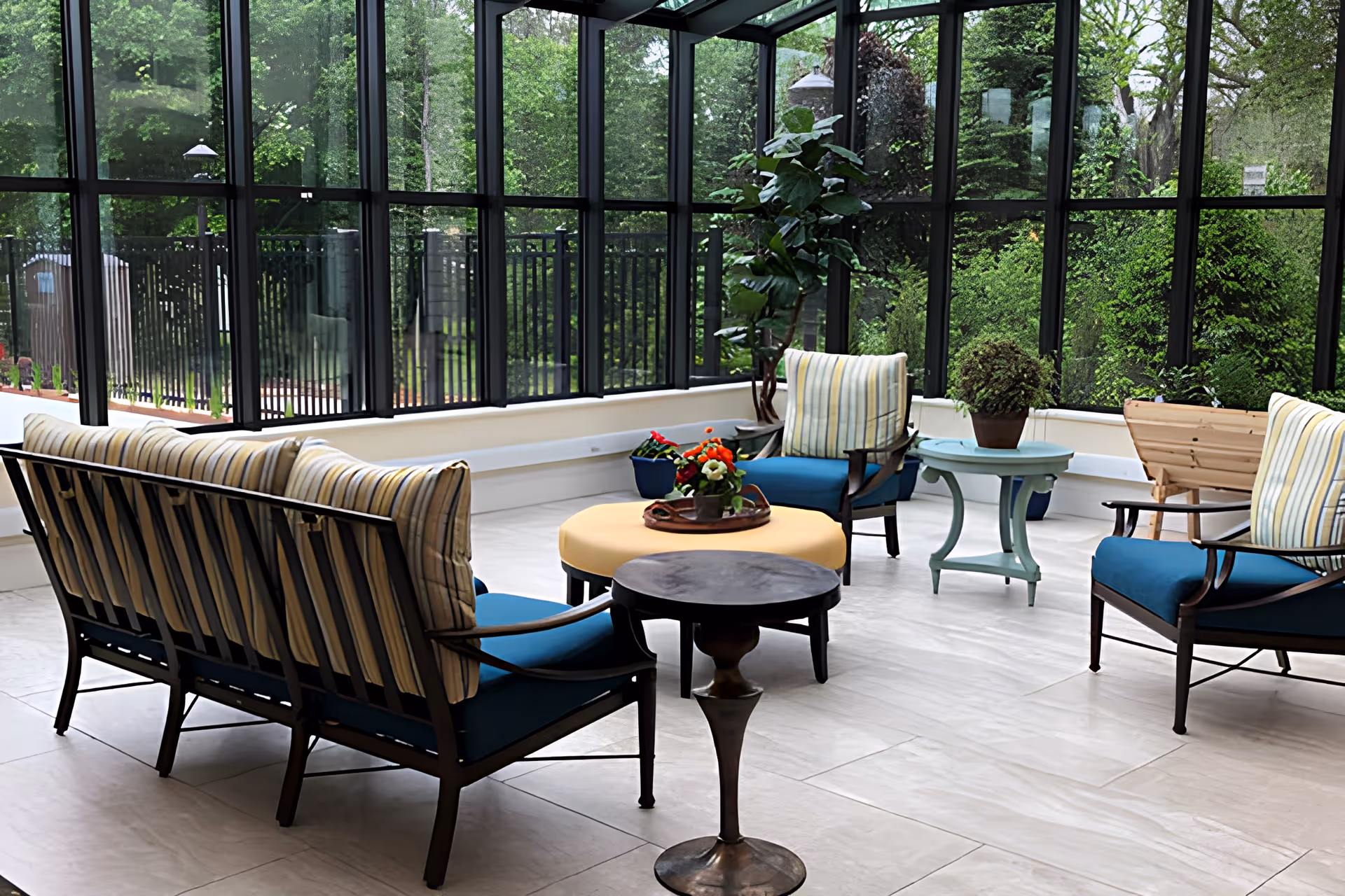 A bright indoor seating area with large floor-to-ceiling windows showing green trees outside. The space has cushioned chairs and a sofa with striped pillows, a round yellow ottoman with a tray of flowers, and small side tables with potted plants.