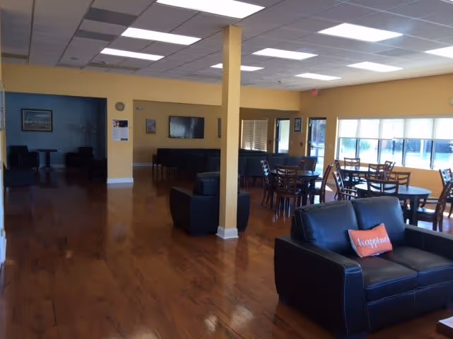 A spacious common area with wooden floors and yellow walls featuring multiple seating arrangements including black leather sofas and wooden tables with chairs. Large windows on the right side allow natural light to fill the room. A pillow with the word 'happiness' is placed on one of the sofas. The ceiling has recessed lighting and a central support column is visible.
