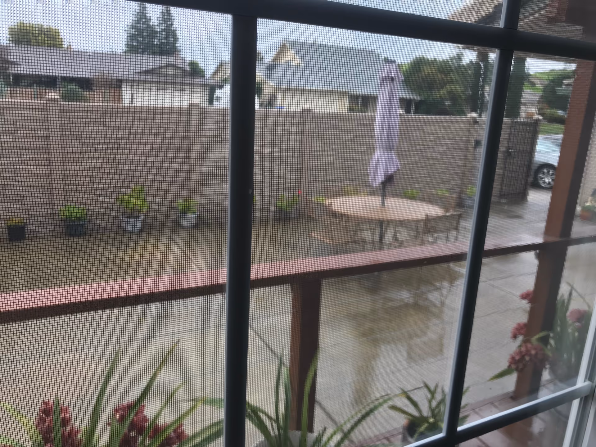 View through a window with a screen showing a fenced outdoor patio area with a round table, chairs, a closed umbrella, and several potted plants along the fence and near the window.