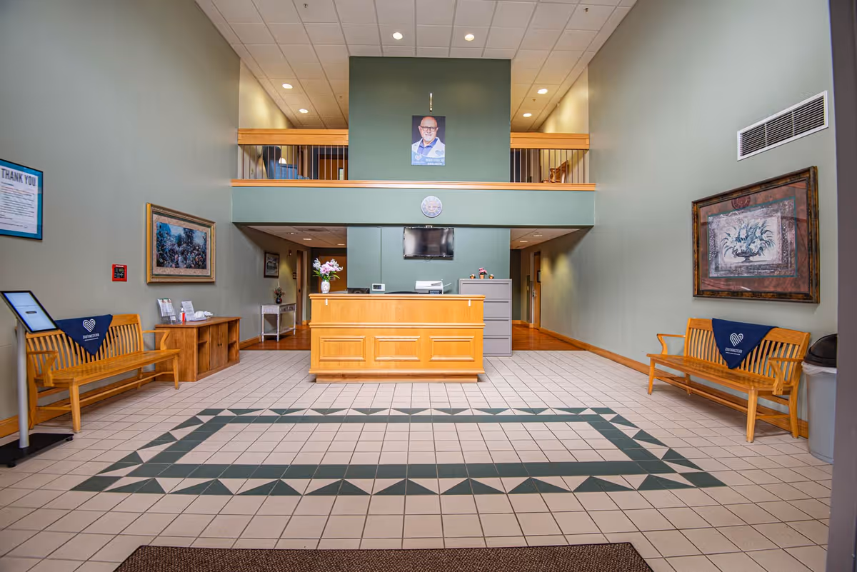 Reception area of Southwestern Nursing and Rehabilitation Center with a wooden front desk in the center, two wooden benches with blue Southwestern Nursing and Rehabilitation Center cloths on either side, framed artwork on the walls, a tiled floor with a green geometric pattern, and a portrait of a man hanging above the desk on a green wall.