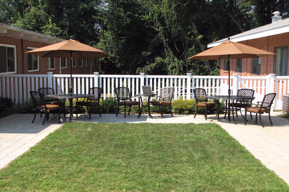 Outdoor patio area with two round tables, each shaded by a large brown umbrella. Each table is surrounded by four metal chairs with brown cushions. The patio is paved with light-colored stones and bordered by a white picket fence. Behind the fence are green bushes and tall trees. The patio is adjacent to a red brick building with several windows.