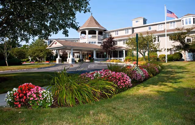 Front entrance of a multi-story senior living building with a covered porte-cochère, landscaped flower beds, and an American flag.