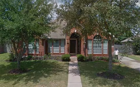 Front exterior view of a single-story brick house with a pathway leading to the front door, flanked by two large windows and surrounded by green grass and trees.