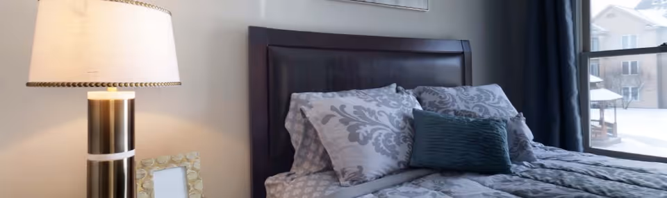 A neatly made bed with patterned bedding and pillows next to a bedside lamp and a window showing buildings outside.