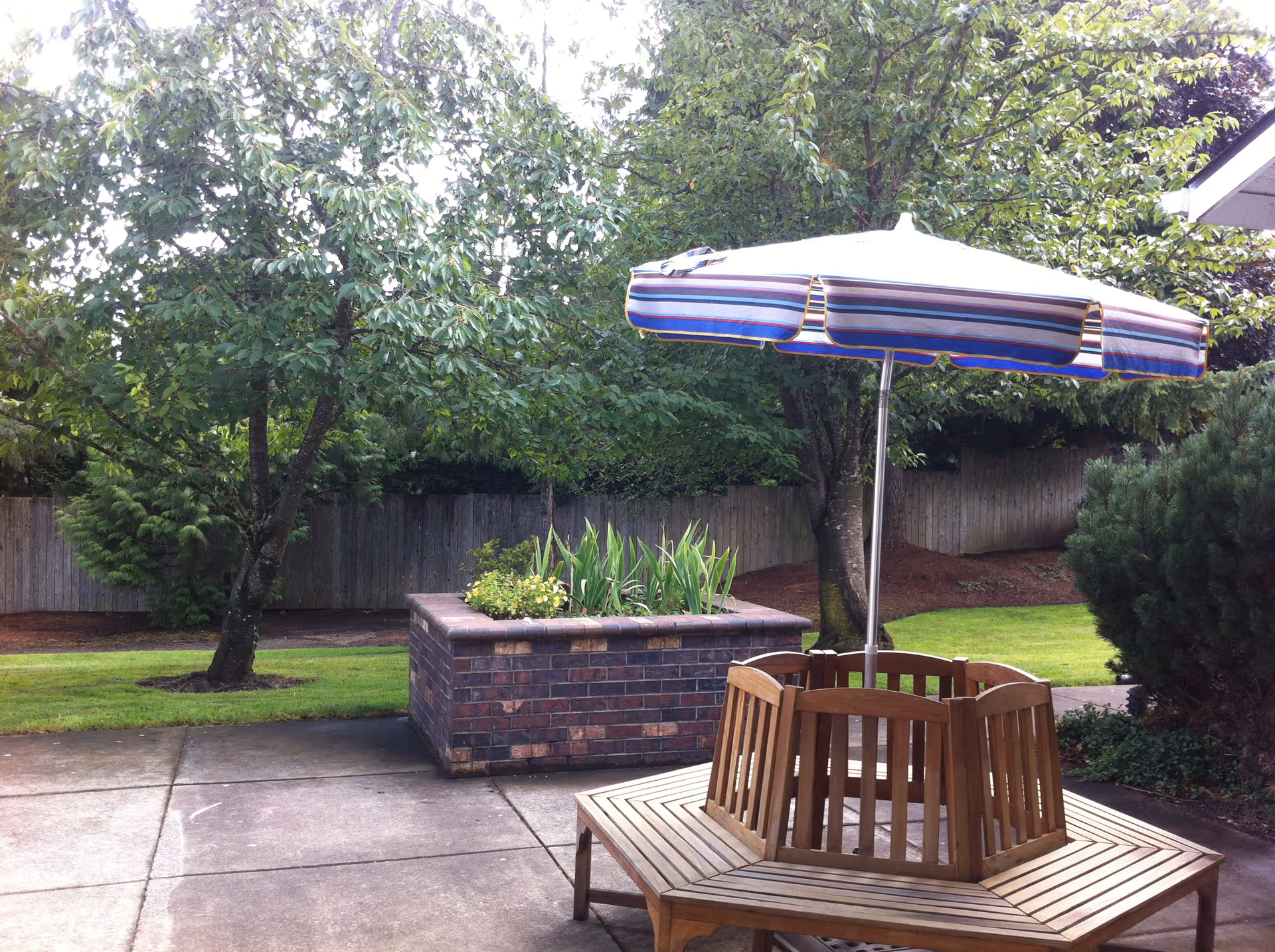 Patio area with a wooden hexagonal bench around a striped umbrella, a brick planter, and trees with a wooden fence in the background.