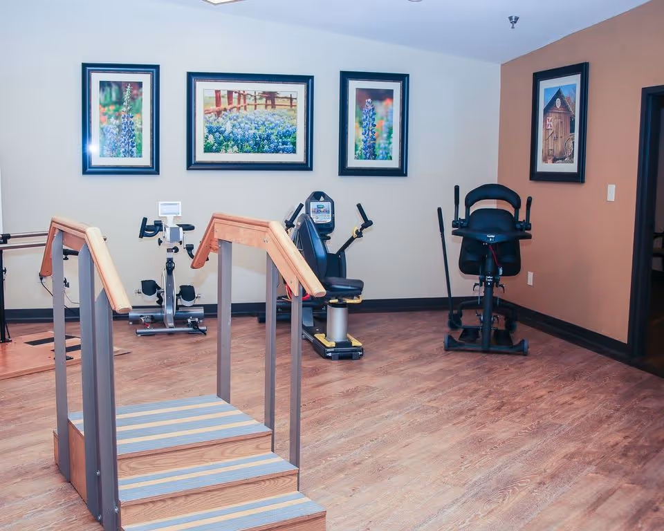 A small exercise room with wooden flooring featuring a set of three-step stairs with handrails, two stationary exercise bikes, and framed pictures of flowers and a barn on the walls.