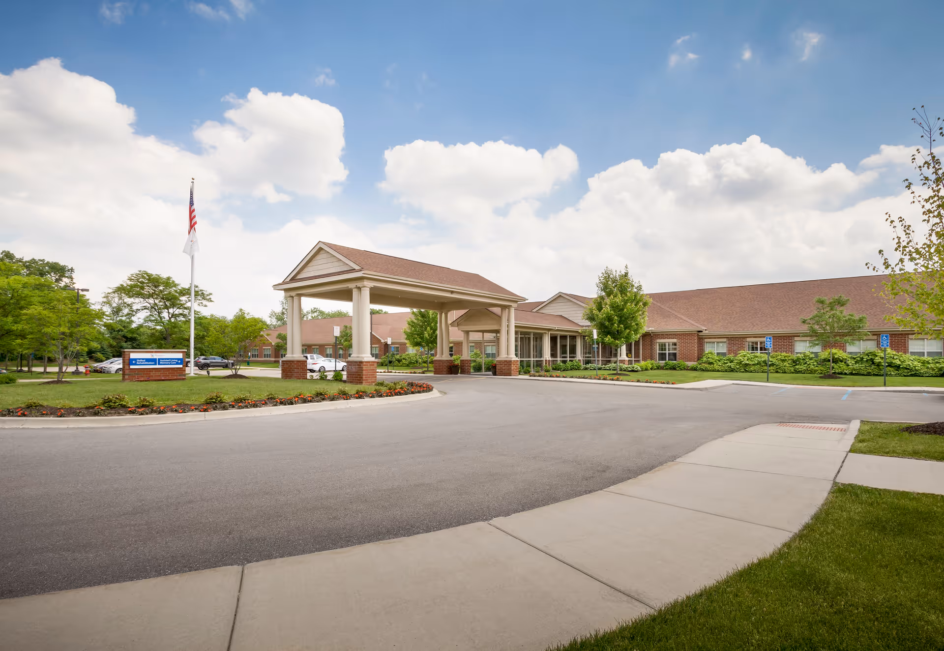Exterior view of Pomeroy Living Rochester Assisted Living and Memory Care facility showing a large covered entrance with columns, a driveway, landscaped flower beds, an American flag, and a clear blue sky with clouds.