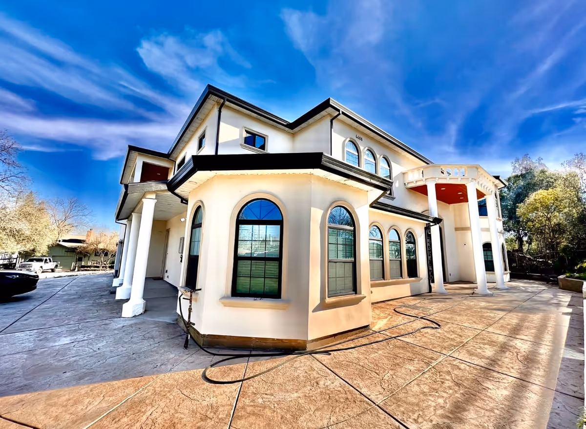 Exterior view of a large two-story building with white walls and multiple arched windows under a bright blue sky with wispy clouds. The building has a covered entrance supported by white columns and a spacious paved area in front. Trees and parked vehicles are visible in the background.