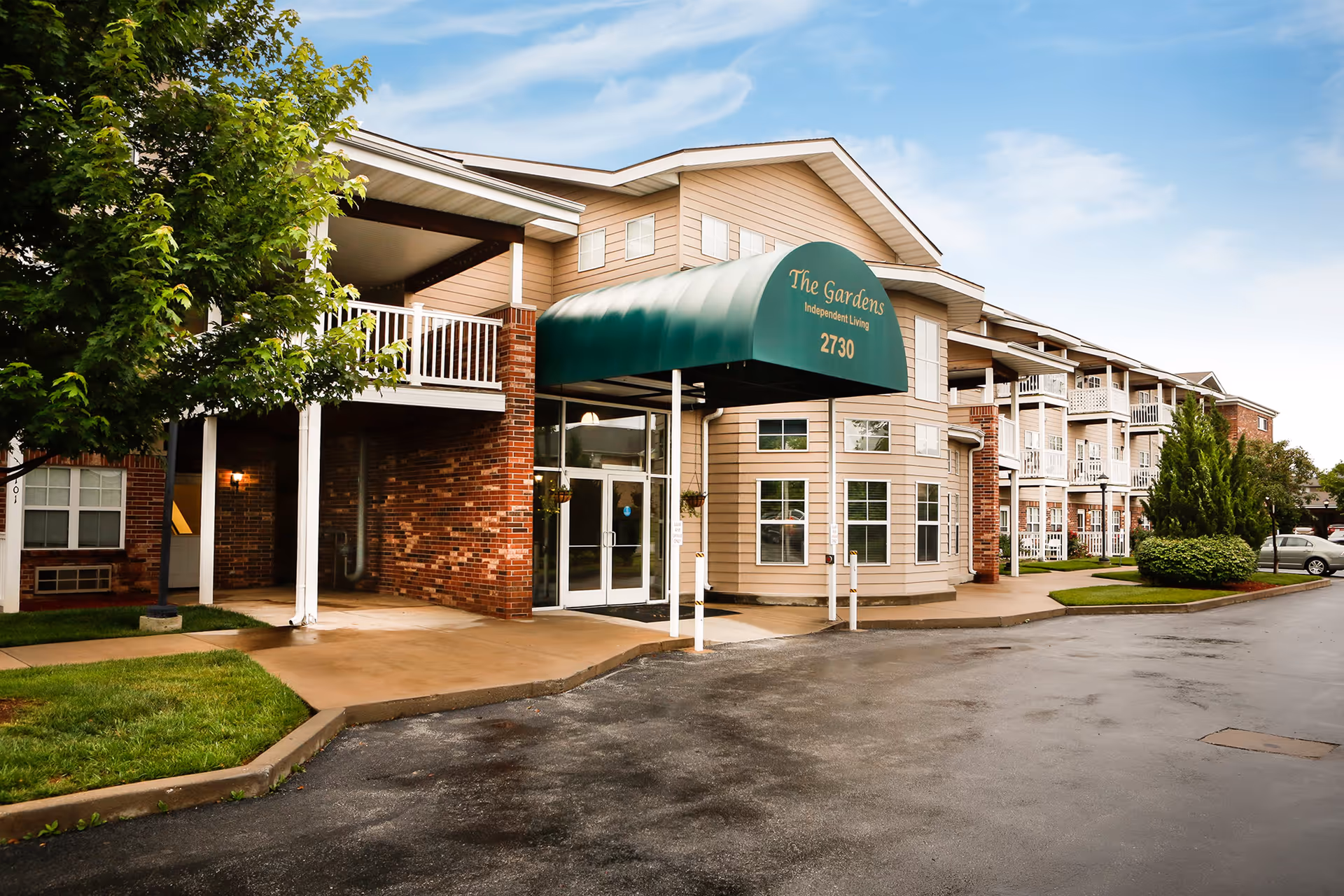Exterior view of The Gardens Independent Living facility showing a multi-story building with balconies, a green awning over the entrance displaying the name and address, surrounded by trees and a paved driveway.