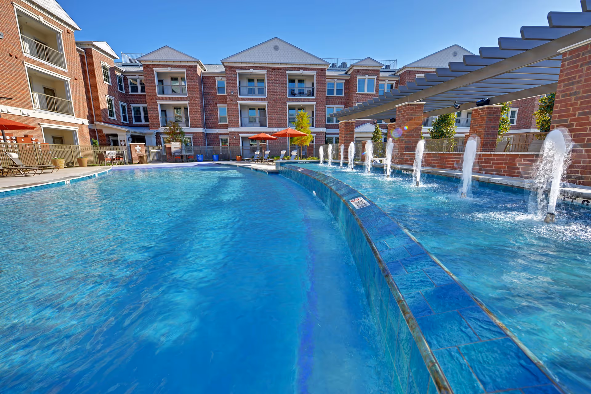 Outdoor swimming pool with water jets and lounge chairs in a courtyard surrounded by a three-story red-brick senior living building.