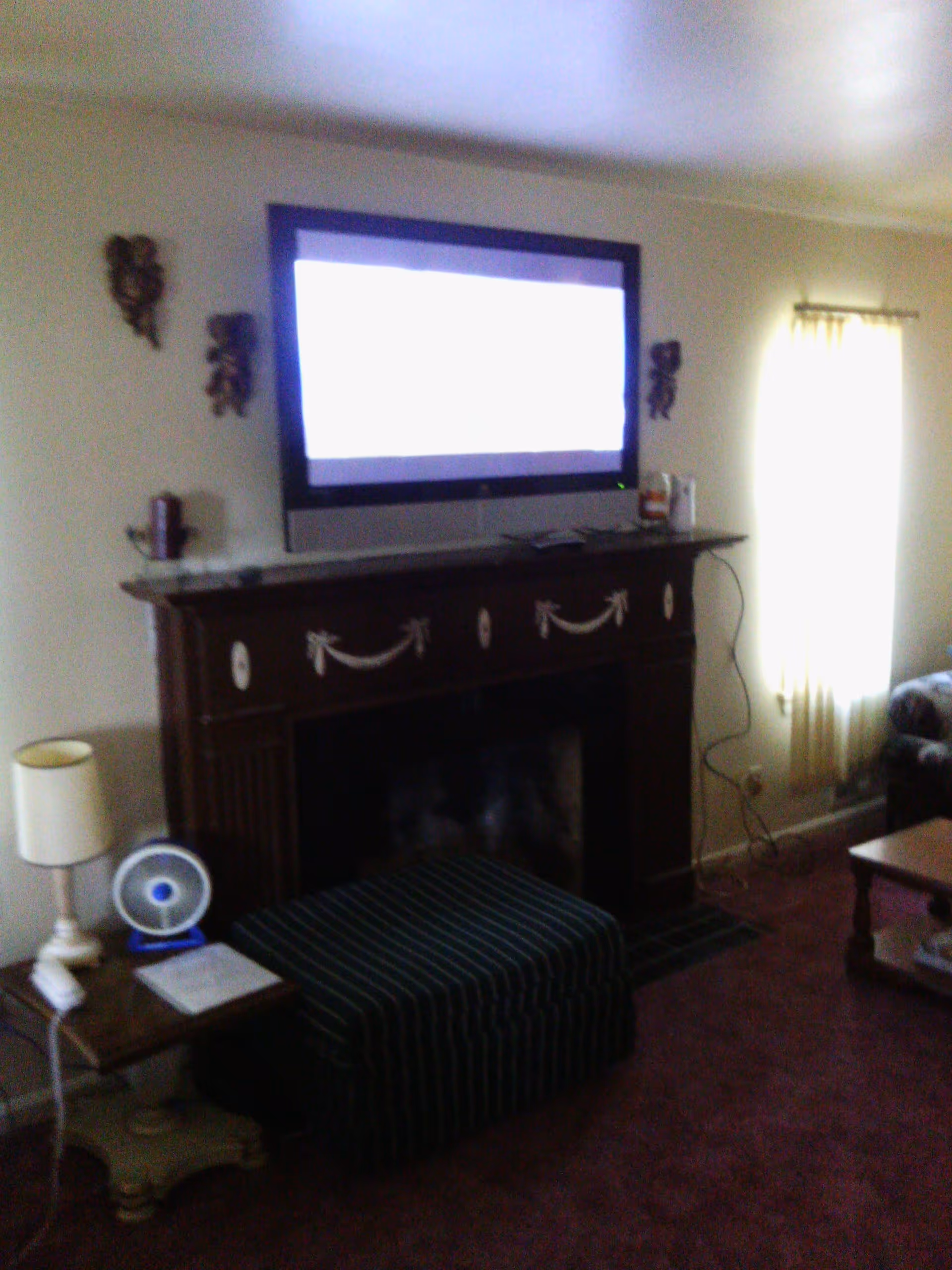 A living room with a mounted flat-screen TV above a decorative dark wooden fireplace. There is a small table with a lamp and a small fan on it to the left, a striped ottoman in front of the fireplace, and a window with light-colored curtains on the right side. Part of a couch and a coffee table are visible on the right side of the image.