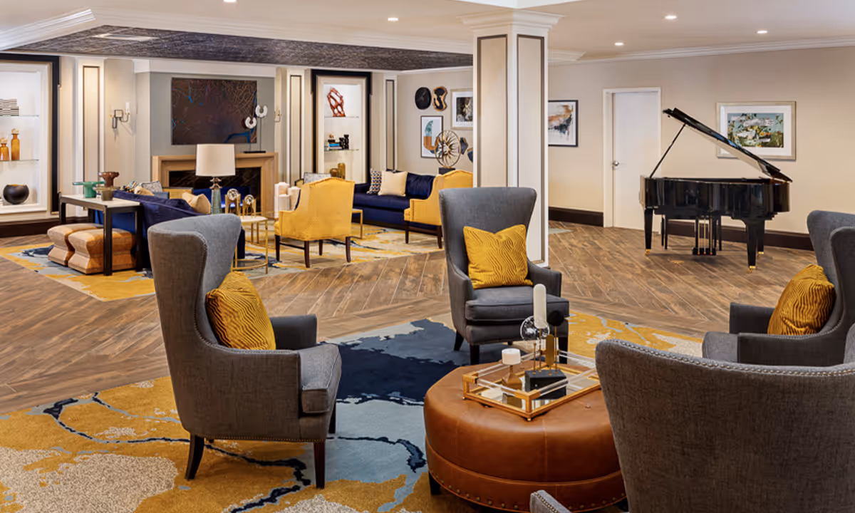A spacious and elegantly decorated living room area in a senior living facility featuring gray armchairs with yellow pillows, a round leather ottoman with decorative items, a grand piano in the background, blue and yellow sofas, and various pieces of wall art and shelving with decorative objects.