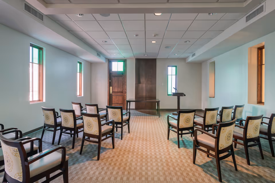 A small conference or meeting room with rows of wooden chairs with patterned cushions facing a wooden door and a podium with a microphone. The room has light-colored walls, three narrow windows, and a beige carpeted floor.