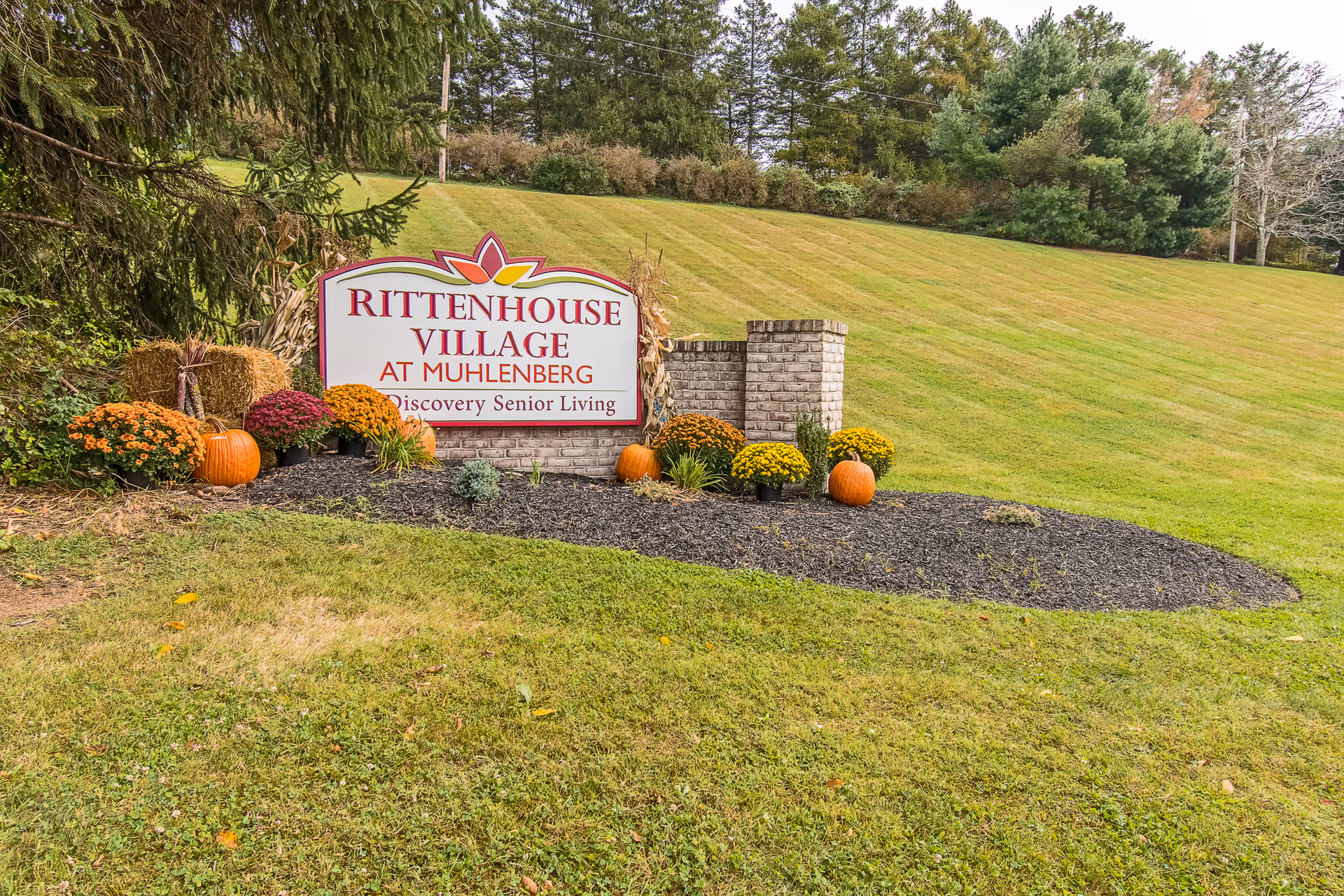 A landscaped outdoor area with a sign that reads 'Rittenhouse Village At Muhlenberg Discovery Senior Living' surrounded by pumpkins, hay bales, and colorful flowers, with a grassy hill and trees in the background.