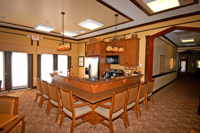 Interior view of a retirement center kitchen and dining area with a large wooden island counter surrounded by chairs. The kitchen features wooden cabinets, a stainless steel refrigerator, microwave, and stove. Decorative hanging lights illuminate the space, and there are large windows with curtains letting in natural light. A hallway extends to the right.