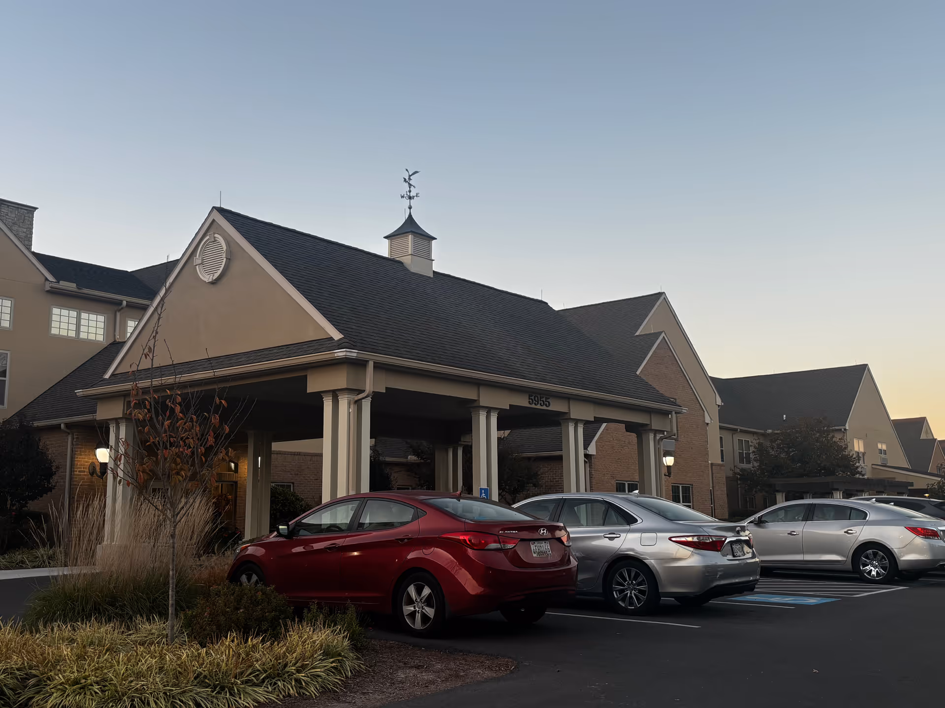 Exterior view of a retirement community building with a covered entrance and several parked cars in front during early evening.