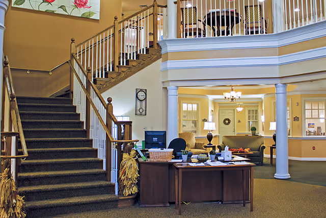 Interior lobby of a senior living facility with a reception desk, seating area, and a carpeted staircase leading to an upper balcony.